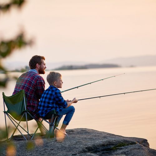 Side view portrait of father and son sitting together on rocks fishing with rods in calm lake waters with landscape of setting sun, both wearing checkered shirts, shot from behind tree