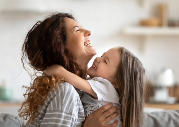 Close up side view overjoyed smiling young mother and daughter hugging and laughing, enjoying tender moment, happy mum and adorable preschool girl kid cuddling, having fun together at home